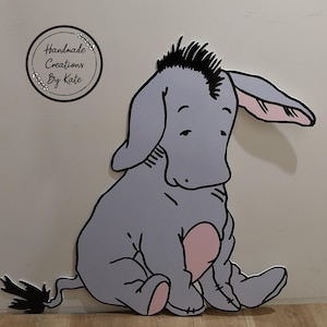 Puede incluir: Un burro de dibujos animados gris y rosa con una melena negra sentado en una superficie de madera. El burro tiene una expresi&oacute;n triste y mira hacia abajo. El texto "Handmade Creations By Kate" es visible en la esquina superior izquierda de la imagen.