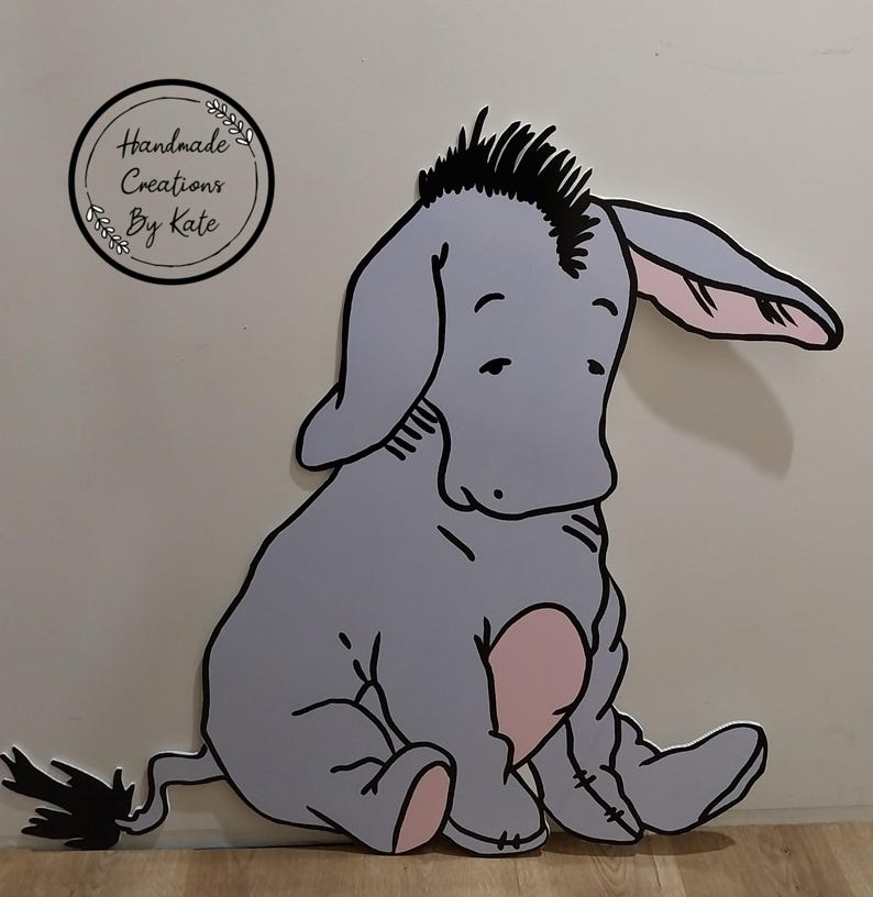 Puede incluir: Un burro de dibujos animados gris y rosa con una melena negra sentado en una superficie de madera. El burro tiene una expresi&oacute;n triste y mira hacia abajo. El texto "Handmade Creations By Kate" es visible en la esquina superior izquierda de la imagen.