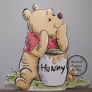 Puede incluir: Una ilustraci&oacute;n de dibujos animados de Winnie de Pooh, un oso de miel, sentado con los brazos cruzados y apoyado en un tarro de miel que dice "Hunny".