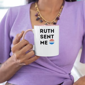 Puede incluir: Taza de cerámica blanca con borde y asa de color rosa. La taza tiene texto negro que dice "Ruth Sent Me" con una pegatina azul y roja que dice "I Dissent".