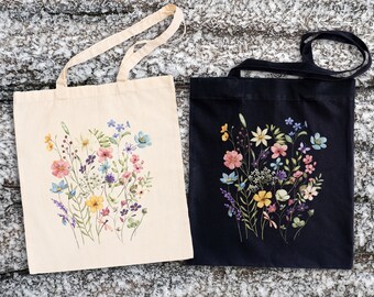 Könnte beinhalten: Zwei Tragetaschen mit Blumendesigns. Eine Tasche ist weiß mit einem bunten Blumendruck, die andere Tasche ist schwarz mit einem ähnlichen Blumendruck in Blautönen, Rosa und Gelb.
