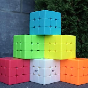 Peut inclure: Une pyramide composée de six cubes de puzzle 3x3 aux couleurs unies. Le cube supérieur est cyan, la rangée centrale présente des cubes vert lime et jaune, et la rangée inférieure se compose de cubes rouge, blanc et orange. Le cube blanc porte un petit texte noir.