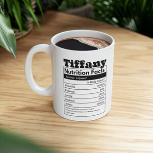 May include: White ceramic mug with the name "Tiffany" and "Nutrition Facts" printed in black. The mug's label lists positive attributes like "Beautiful," "Patience," and "Loving," with percentage values. The mug is filled with dark coffee.