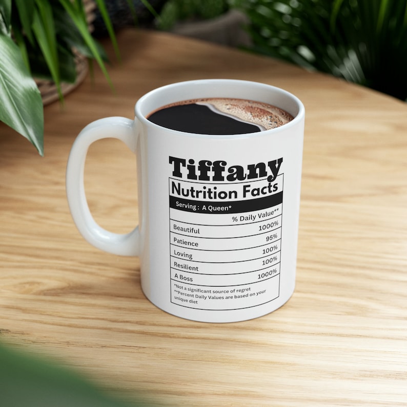 May include: White ceramic mug with the name "Tiffany" and "Nutrition Facts" printed in black. The mug's label lists positive attributes like "Beautiful," "Patience," and "Loving," with percentage values. The mug is filled with dark coffee.
