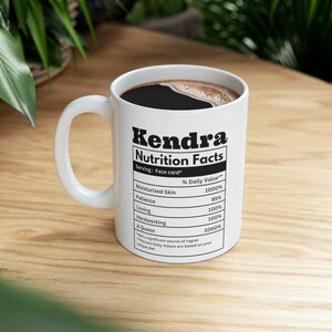 May include: White ceramic mug with the name "Kendra" and "Nutrition Facts" printed in black. The mug's label lists positive attributes like "Moisturized Skin" and "A Queen" with corresponding percentage values. The mug is filled with dark liquid.