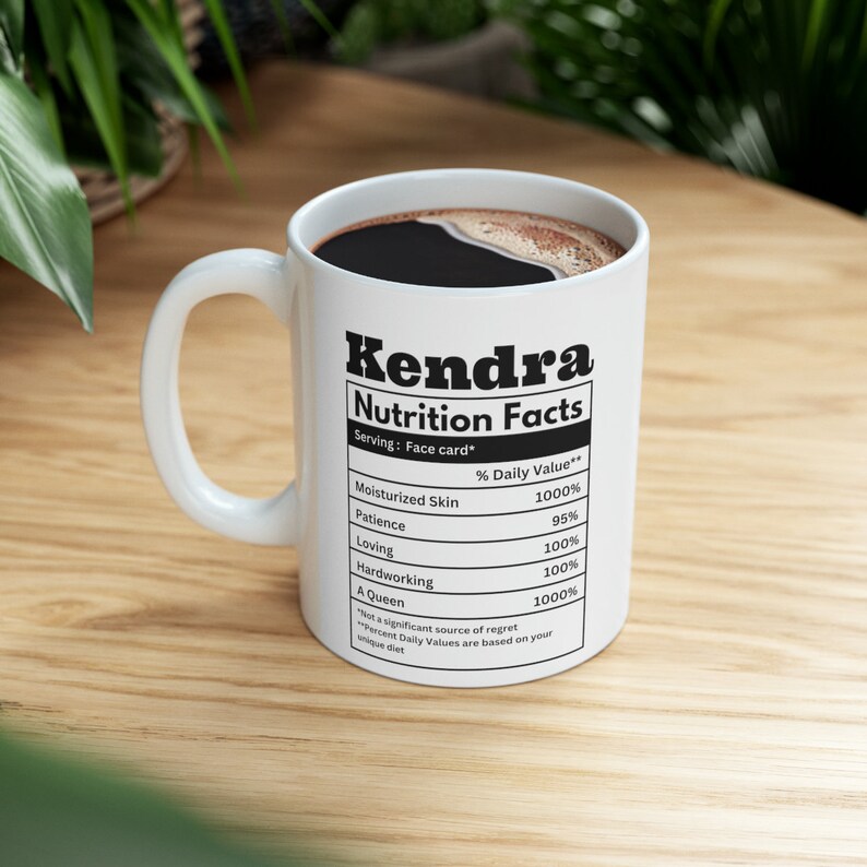 May include: White ceramic mug with the name "Kendra" and "Nutrition Facts" printed in black. The mug's label lists positive attributes like "Moisturized Skin" and "A Queen" with corresponding percentage values. The mug is filled with dark liquid.