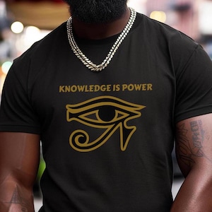 May include: Black t-shirt with the text "KNOWLEDGE IS POWER" in gold lettering above a gold Eye of Horus symbol. The shirt is worn by a person with a gold chain necklace.