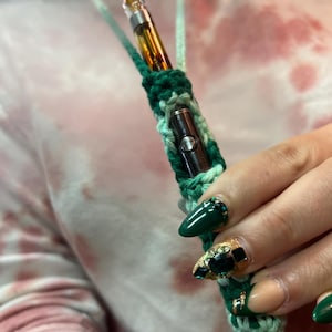 May include: A green and white crocheted necklace with a small, clear glass vape pen inside. The necklace is being held by a hand with green and gold nail polish.