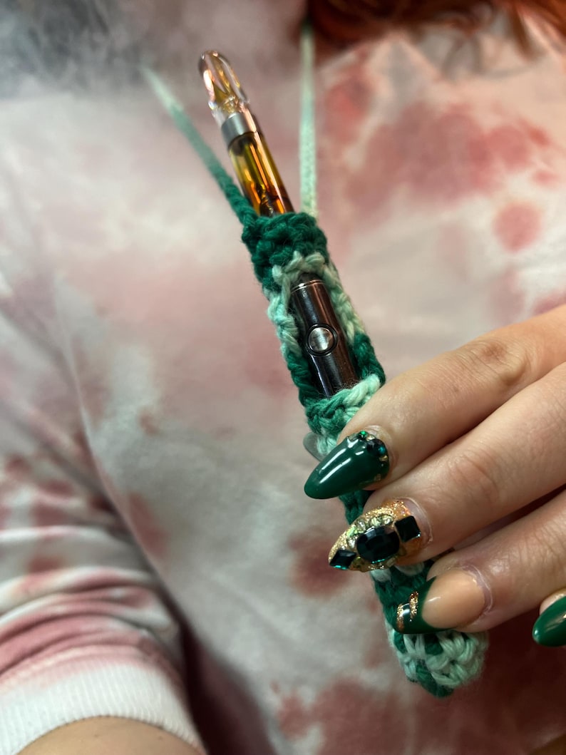 May include: A green and white crocheted necklace with a small, clear glass vape pen inside. The necklace is being held by a hand with green and gold nail polish.