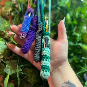 May include: Four crocheted vape pen cases in different colors, including purple, blue, green, and gray. The cases are held in a hand against a green leafy background.