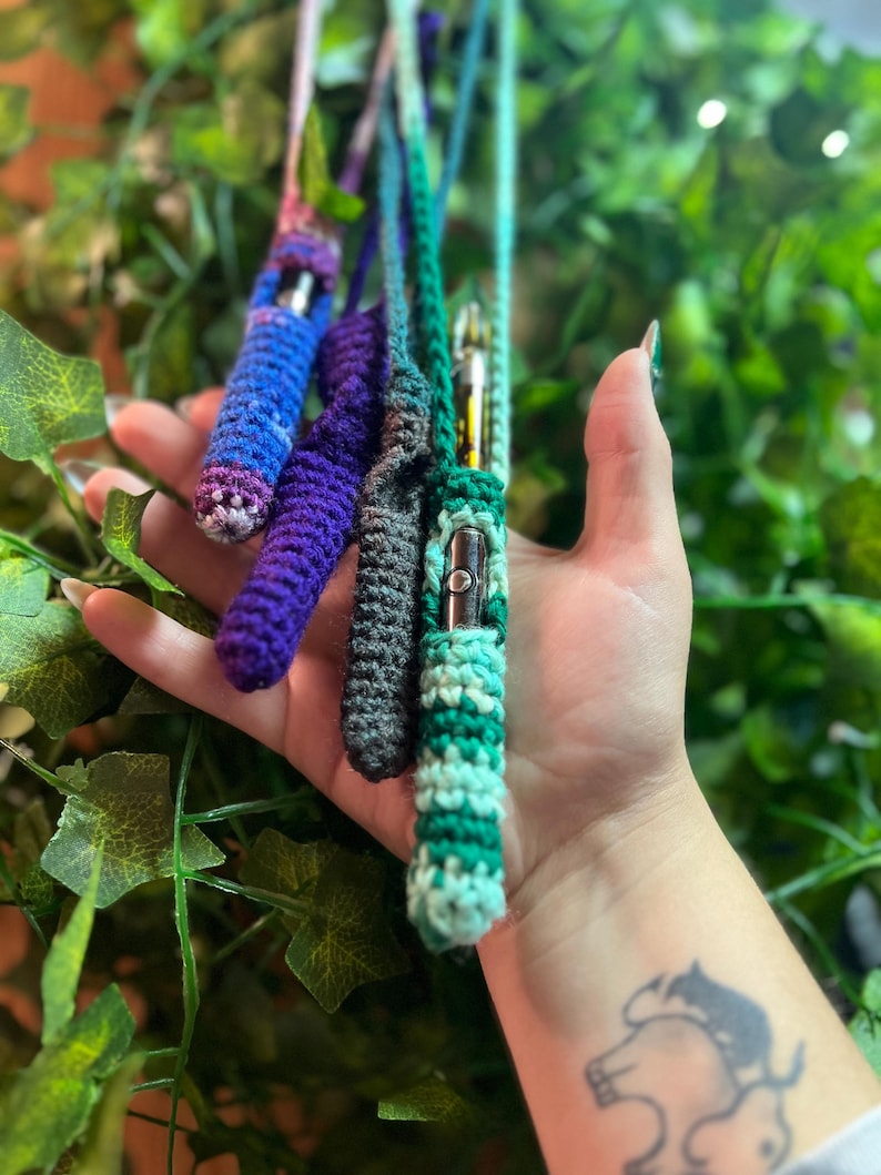 May include: Four crocheted vape pen cases in different colors, including purple, blue, green, and gray. The cases are held in a hand against a green leafy background.