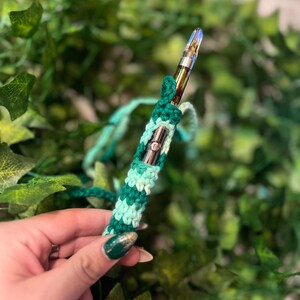 May include: A green and white crocheted vape pen sleeve. The sleeve is made with a simple stitch pattern and has a light green and dark green color scheme.