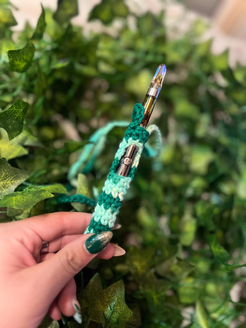 May include: A green and white crocheted vape pen sleeve. The sleeve is made with a simple stitch pattern and has a light green and dark green color scheme.