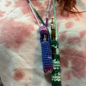 May include: A crocheted necklace with two pouches for holding vape cartridges. The pouches are green, purple, and blue.