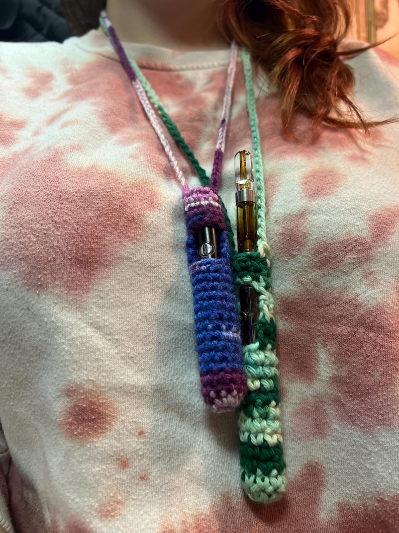 May include: A crocheted necklace with two pouches for holding vape cartridges. The pouches are green, purple, and blue.