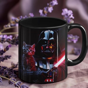 May include: A black ceramic mug with a graphic of Darth Vader and a kitten. The kitten is wearing a red collar and is looking at the viewer. Darth Vader is holding a red lightsaber. The mug has a black handle.