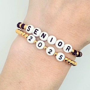 May include: Two beaded bracelets with black and gold beads. The top bracelet spells out "SENIOR" in white beads with black lettering. The bottom bracelet spells out "2025" in white beads with black lettering.