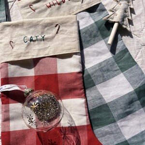 May include: Two fabric Christmas stockings featuring a red and white buffalo check pattern and a green and white buffalo check pattern. Each stocking has a cream-coloured cuff embroidered with the names CATY and WILBO and small candy canes.