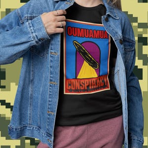 Könnte beinhalten: Schwarzes T-Shirt mit dem Text "Oumuamua Conspiracy" in einem Grafikdesign. Das Design zeigt eine stilisierte Darstellung des interstellaren Objekts vor blauem und violettem Hintergrund mit gelben Strahlen. Das T-Shirt wird mit einer Jeansjacke getragen.