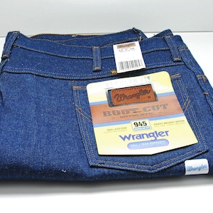 Op de afbeelding: Een paar blauwe Wrangler 945 Boot Cut jeans van denim met een bruin leren patch waarop "Wrangler Boot Cut" staat en een wit label met de tekst "Wrangler 945" en "Full-Tear Warranty".