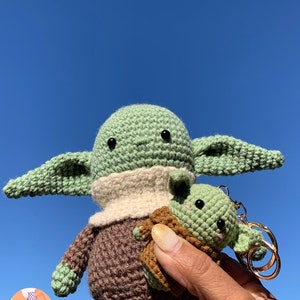 May include: Handmade crochet figures of a character with large ears, green skin, and a brown robe. One figure is larger, and the other is a keychain. The background is a clear blue sky. The figures are made by Jani Crochet.