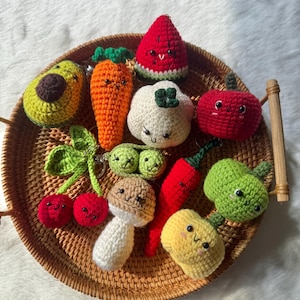 May include: A collection of colorful, crocheted food-themed keychains, including an avocado, carrot, watermelon, cauliflower, tomato, cherries, peas, mushroom, chili pepper, and a pear. Each keychain has a smiling face and is arranged on a woven tray.