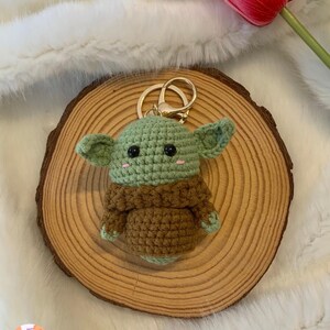 May include: A handmade crochet keychain featuring a green and brown character with large ears and black eyes. The keychain is attached to a gold-colored ring. The character is placed on a wooden slice, with a red tulip in the background.