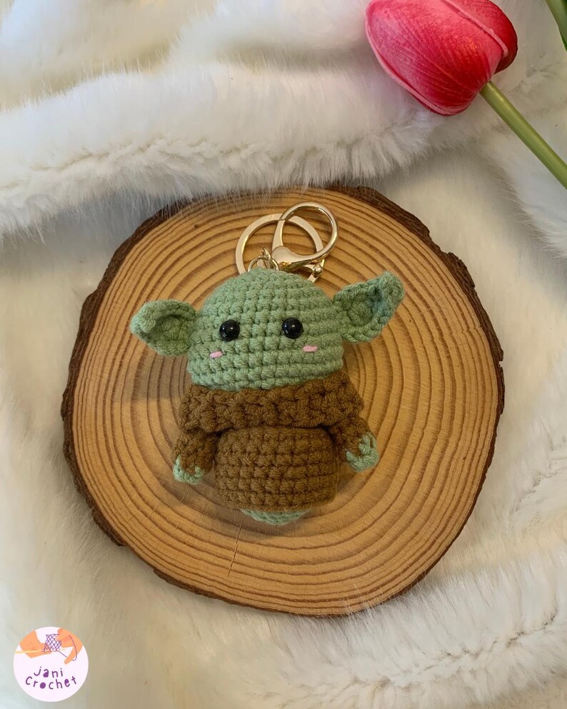 May include: A handmade crochet keychain featuring a green and brown character with large ears and black eyes. The keychain is attached to a gold-colored ring. The character is placed on a wooden slice, with a red tulip in the background.