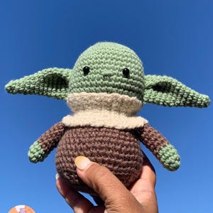 May include: A handmade crochet doll in the likeness of a popular character. The doll features a green head and ears, a tan collar, and a brown body and arms. The doll is held against a bright blue sky.