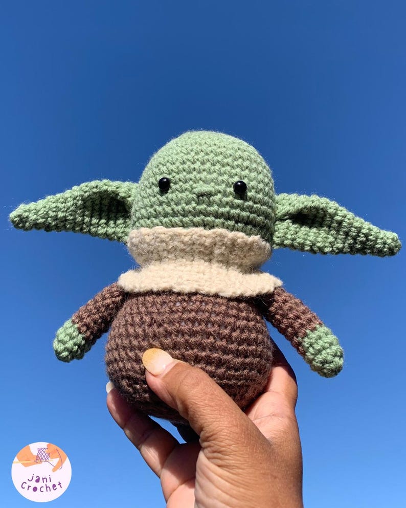 May include: A handmade crochet doll in the likeness of a popular character. The doll features a green head and ears, a tan collar, and a brown body and arms. The doll is held against a bright blue sky.