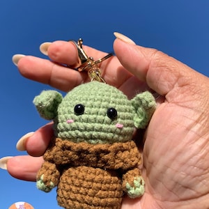 May include: A handmade crochet keychain featuring a green and brown character with large black eyes and small pink cheeks. The keychain has a gold-colored clasp and ring. The character is held against a bright blue sky.