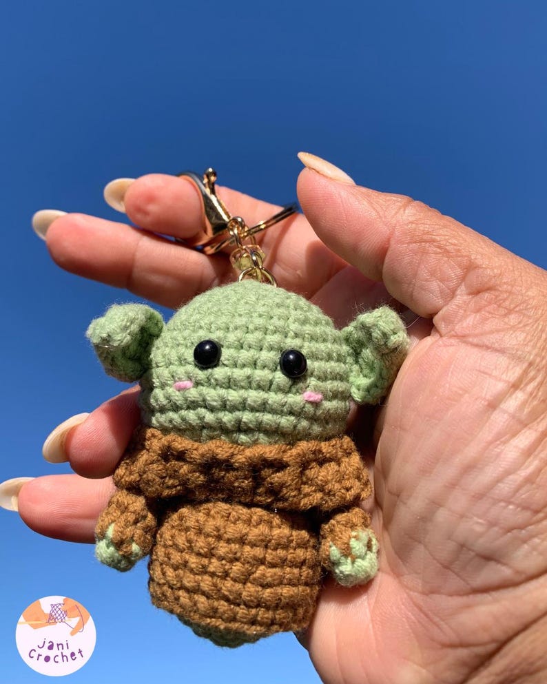 May include: A handmade crochet keychain featuring a green and brown character with large black eyes and small pink cheeks. The keychain has a gold-colored clasp and ring. The character is held against a bright blue sky.
