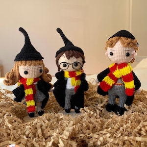 Könnte beinhalten: Drei gehäkelte Puppen von Charakteren aus einer bekannten Fantasy-Serie. Jede Puppe trägt einen schwarzen Hut, einen rot-gelben Schal und eine schwarze Robe. Die Puppen stehen auf geschreddertem Papier.