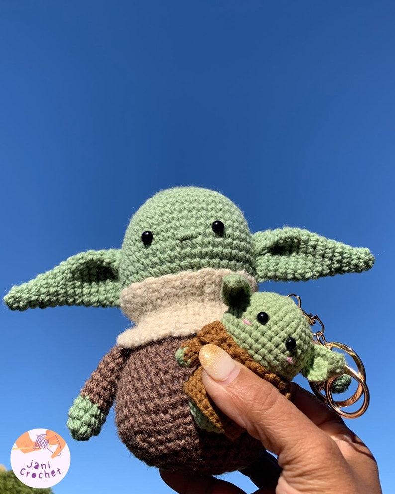 May include: Handmade crochet figures of a character with large ears, green skin, and a brown robe. One figure is larger, and the other is a keychain. The background is a clear blue sky. The figures are made by Jani Crochet.