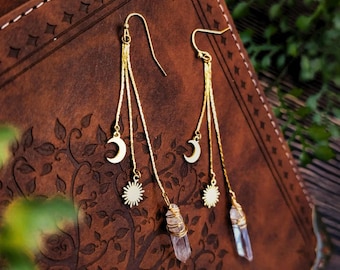 May include: Gold-tone drop earrings with a crescent moon, sun, and a wrapped crystal pendant. The earrings hang from a gold hook and are displayed on a brown, textured surface. The celestial-themed jewellery has a bohemian aesthetic.