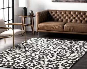 Pode incluir: Tapete retangular com estampa de leopardo em preto e branco. O tapete está colocado sobre um piso de madeira escura. Um sofá de couro marrom e uma cadeira são visíveis ao fundo.