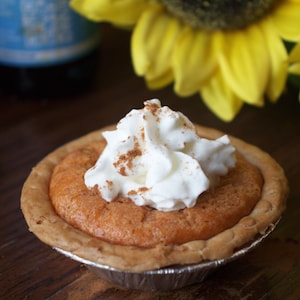 May include: A single-serving pumpkin pie with a golden crust, topped with whipped cream and a sprinkle of cinnamon. The pie sits in a silver foil container, with a yellow sunflower in the background.