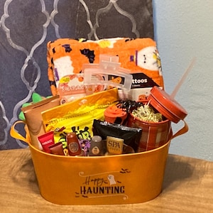May include: An orange metal oval basket with the text Happy Haunting and a ghost graphic, filled with a Halloween-themed plush blanket, a red lidded tumbler with a straw, various small skincare products, and a clear plastic hair claw clip.