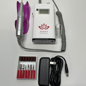 May include: A white nail drill with a digital display and the text "Phalla's Nails & Spa LLC". Includes a silver handpiece, a purple holder, a set of drill bits, and a black power adapter.