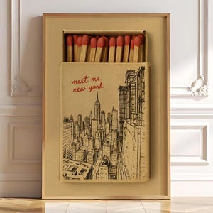 Puede incluir: Obra de arte enmarcada con un dise&ntilde;o de caja de cerillas que representa el horizonte de la ciudad de Nueva York. La caja de cerillas est&aacute; abierta, revelando cerillas con puntas rojas. El texto "meet me new york" est&aacute; escrito en rojo.