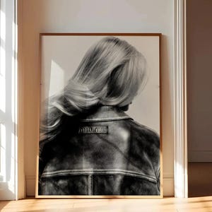 May include: A framed black and white photograph depicting the back view of a person with long hair wearing a dark leather jacket. The jacket features a visible collar, horizontal seam detailing across the back, and the text Miu Miu on a label.