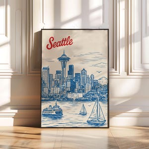 May include: A framed art print featuring a blue and white illustration of the Seattle skyline, including the Space Needle, sailboats, and a ferry. The word "Seattle" is written in red script at the top.