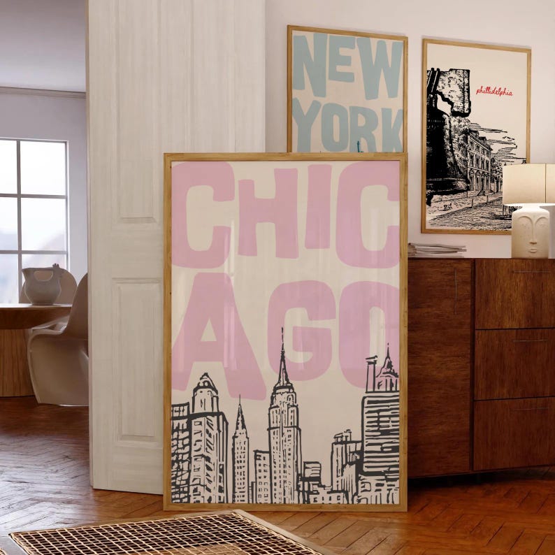 May include: A framed art print featuring the word "CHICAGO" in pink block letters over a black and white skyline illustration. Two other framed prints are visible, one with "NEW YORK" in blue and another with a Philadelphia scene.
