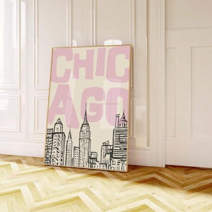 May include: A framed art print featuring the word "CHICAGO" in pink block letters over a black and white skyline illustration. The artwork is displayed against a cream background, with a light wood frame.