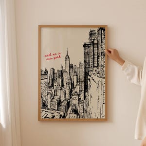 May include: A framed black and white sketch of the New York City skyline, including the Empire State Building. The words "meet me in new york" are written in red in the upper left corner. The frame is made of light brown wood.