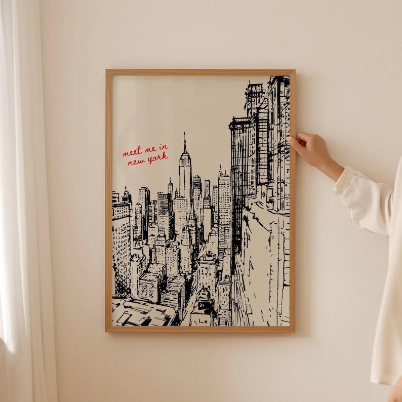 May include: A framed black and white sketch of the New York City skyline, including the Empire State Building. The words "meet me in new york" are written in red in the upper left corner. The frame is made of light brown wood.