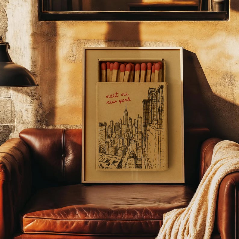 Puede incluir: Una obra de arte enmarcada con un dise&ntilde;o de caja de cerillas que muestra una ilustraci&oacute;n del horizonte de la ciudad de Nueva York. La caja de cerillas contiene cerillas con puntas rojas. El texto "meet me new york" est&aacute; escrito en rojo. La obra se exhibe en un sof&aacute; de cuero marr&oacute;n.