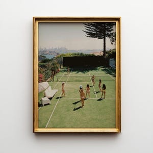 以下が含まれることがあります： テニスコートのシーンの額入りプリント。水着姿の数人が緑のコートでテニスをしています。背景には街並み、木、白いソファとパラソルが見えます。