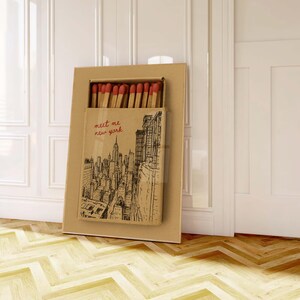 Puede incluir: Una caja de cerillas grande con una ilustraci&oacute;n del horizonte de la ciudad de Nueva York y el texto "meet me new york". La caja de cerillas est&aacute; abierta, revelando cerillas con puntas rojas. La caja es marr&oacute;n y rectangular.