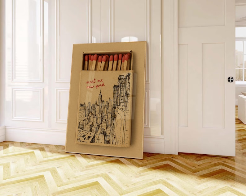 Puede incluir: Una caja de cerillas grande con una ilustraci&oacute;n del horizonte de la ciudad de Nueva York y el texto "meet me new york". La caja de cerillas est&aacute; abierta, revelando cerillas con puntas rojas. La caja es marr&oacute;n y rectangular.